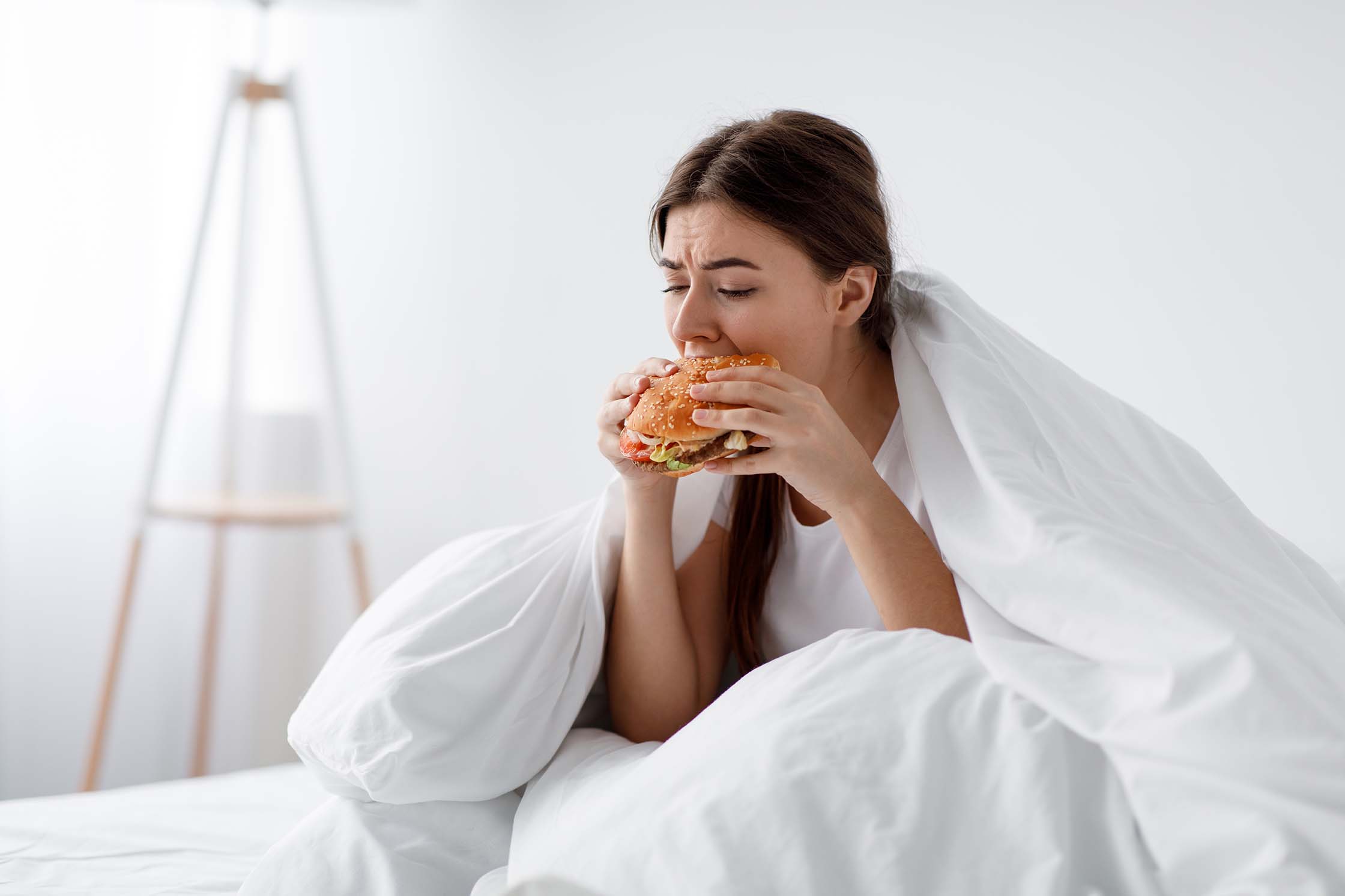Hungry after diet, starving stressed caucasian millennial woman eating burger in bed at home alone. Unhealthy overeating lifestyle, depression, psychological and mental problems. Eating too much
